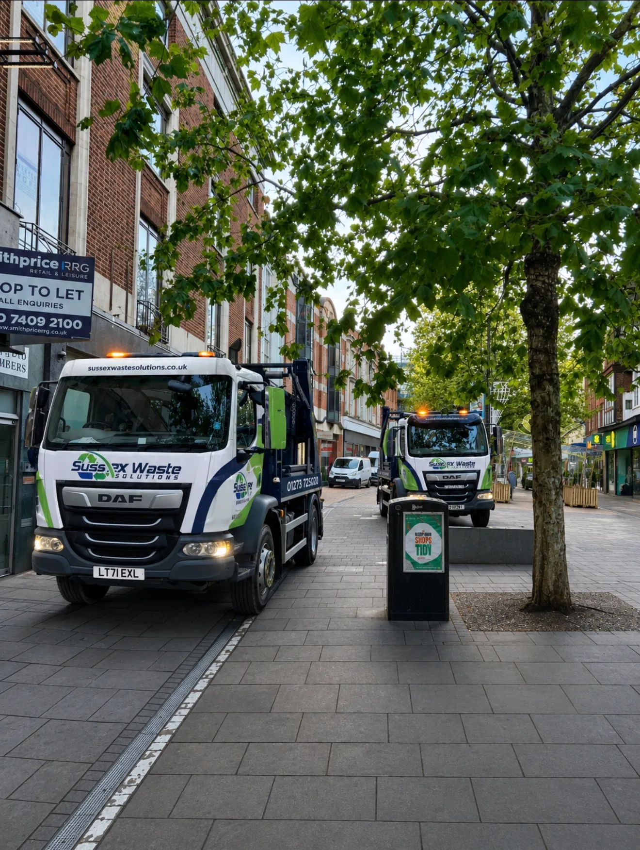 Two Sussex Waste Solutions trucks parked on a city street with tree and trash bin nearby.